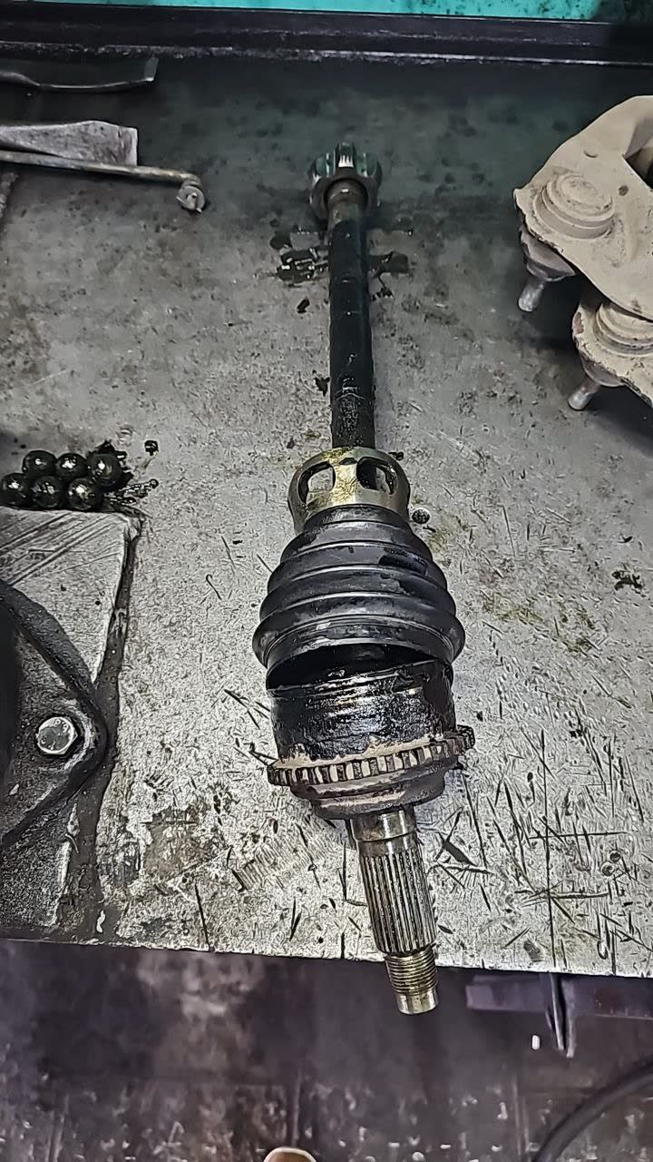 Removed drive shaft on a workbench showing the outer CV joint with its torn/cracked rubber dust boot, splined shaft end and loose bearing balls visible nearby, plus a disassembled brake caliper in the background