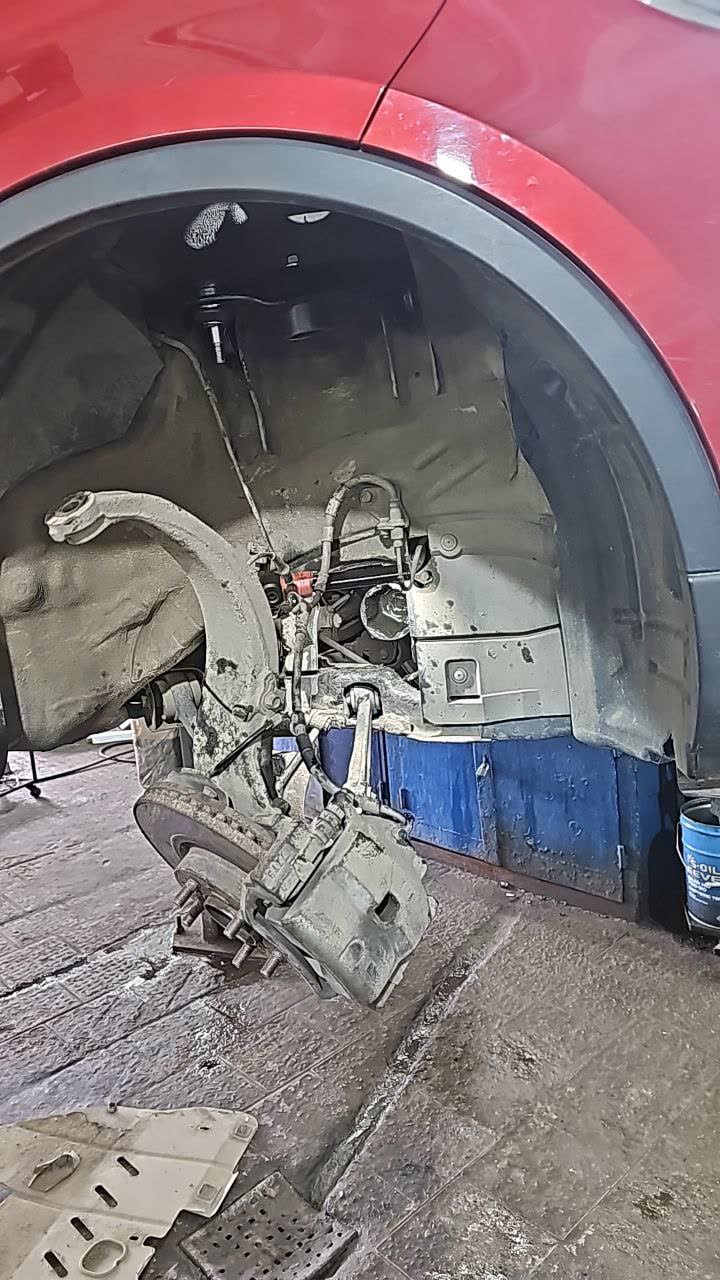 Front wheel removed on a red X80, showing the front suspension/brake assembly (control arm, brake disc, caliper hanging loose) with the driveshaft area exposed for the CV boot repair