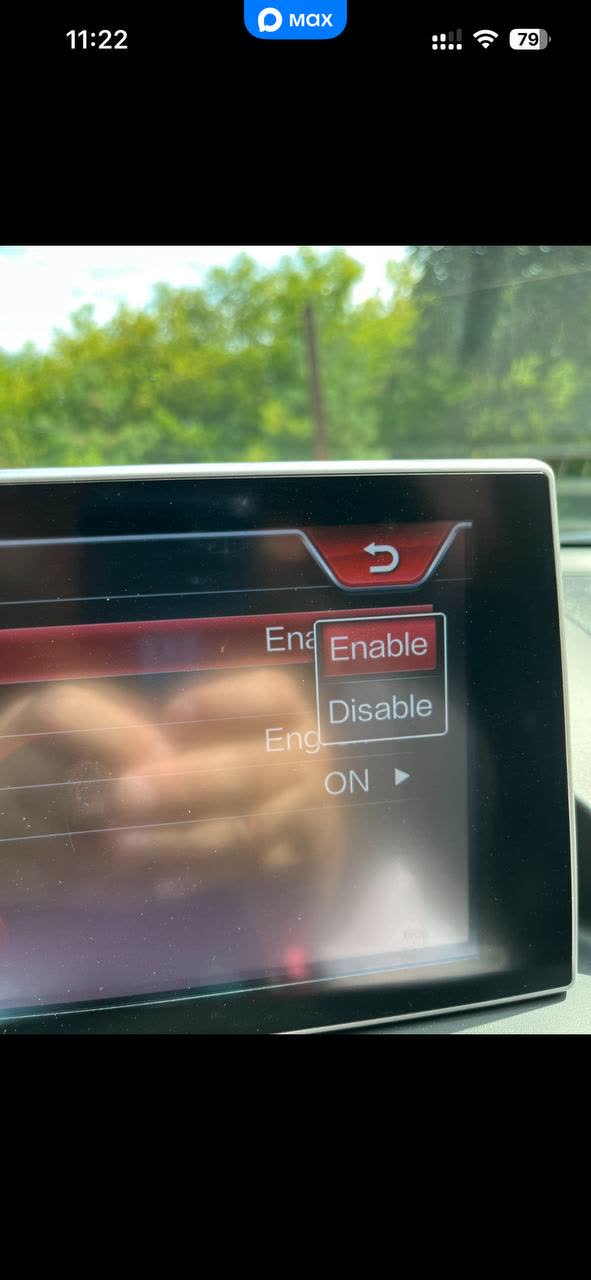Photo of the infotainment touchscreen showing a settings submenu with an "Enable / Disable" toggle (Enable highlighted) and an "ON ▶" row beneath a partly hand-obscured setting label; this is the screen used to allow video playback to continue while the car is in motion.