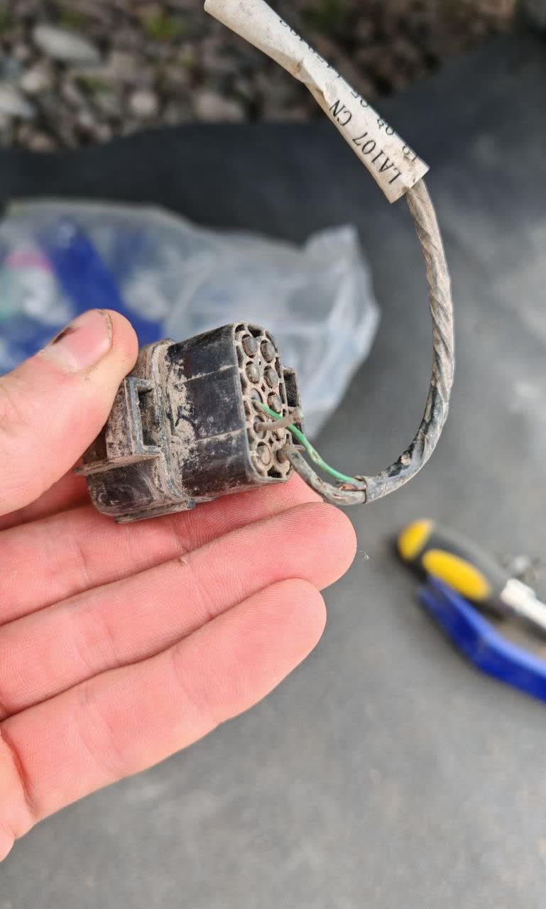 A burnt/degraded multi-pin connector housing (tagged "LA107 CN") pulled from the harness, held in hand next to pliers — only a couple of thin wires (one green) remain intact, illustrating the kind of connector damage found during a DRL wire-chafe repair